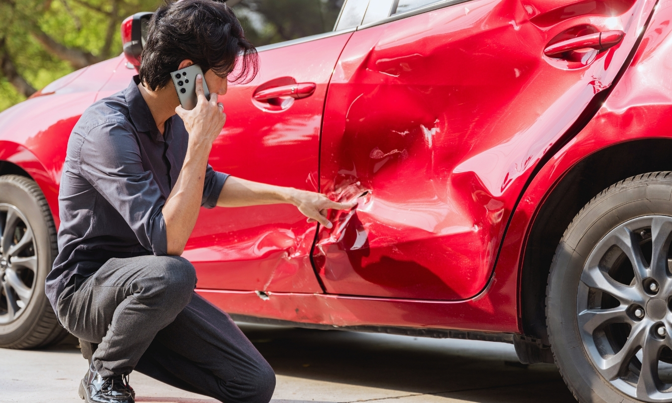 man crouched and on the phone, looking at dented car door